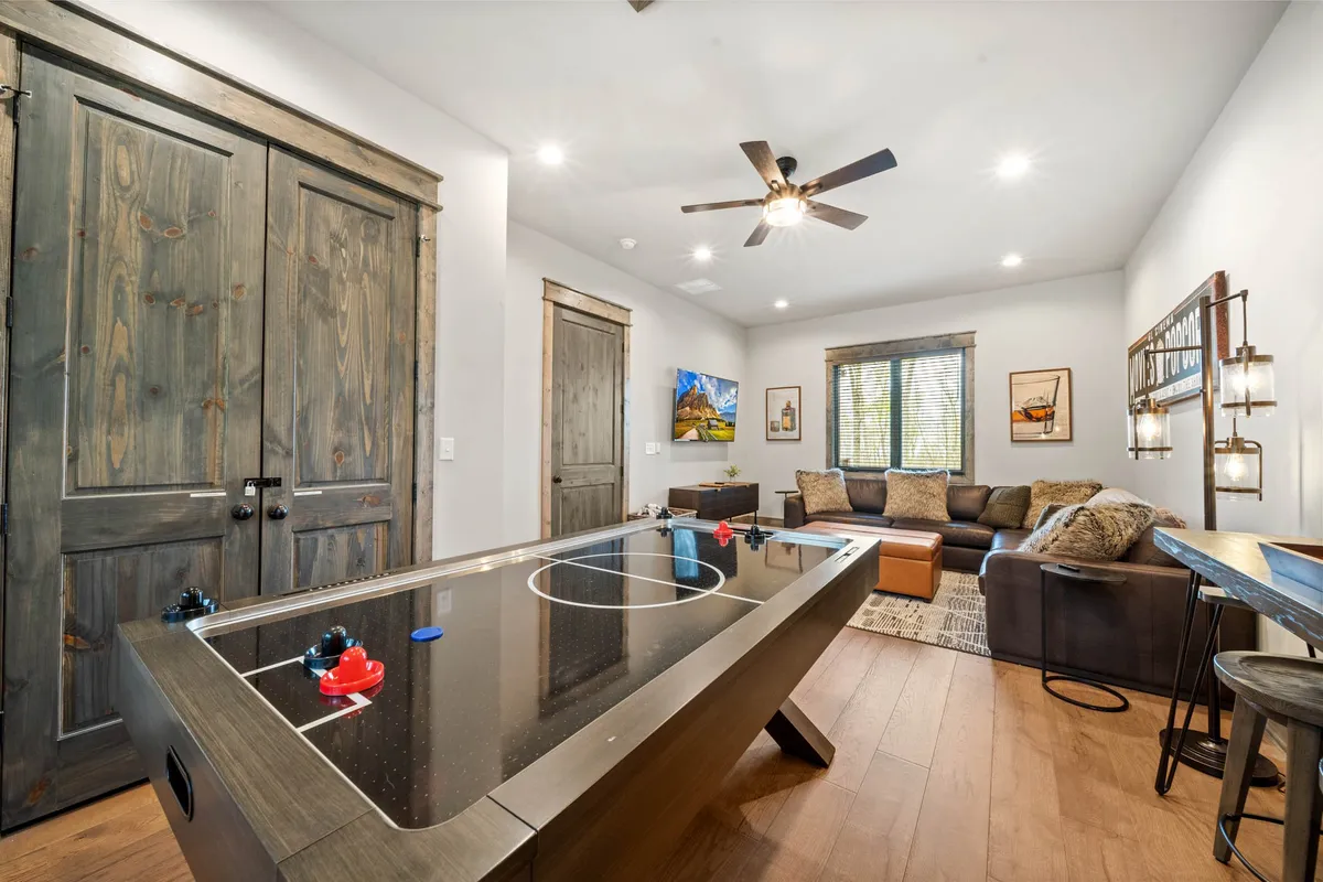 The image shows a lively game room with an air hockey table in the foreground, a comfortable sectional sofa along one wall, stylish artwork, and large wooden doors. People are engaged in activities such as playing air hockey, relaxing on the sofa, or watching TV, giving the space a fun, communal vibe.
