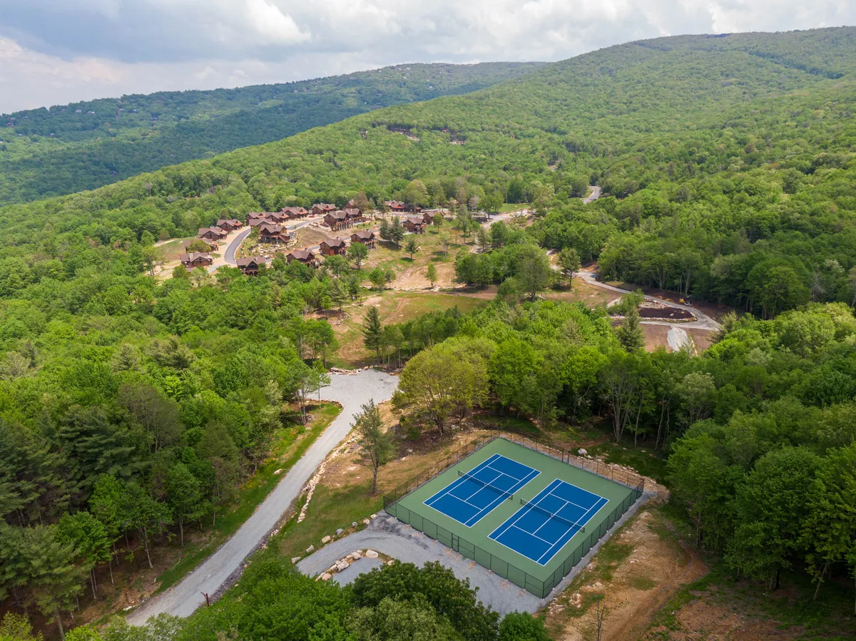 A wide angle shows tennis courts surrounded by rustic wooden cabins and a dramatic mountain backdrop. The setting conveys both active recreation and community spirit, as well as easy access to outdoor sports within the neighborhood.