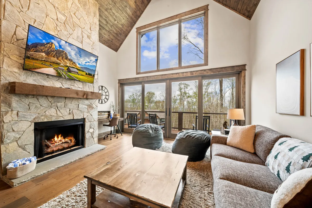 An inviting living room with a stone fireplace as the centerpiece, comfortable sofas, large windows with nature views, a wooden coffee table, and a flatscreen TV displaying a landscape image. The setting beckons for relaxation, storytelling, and movie nights.