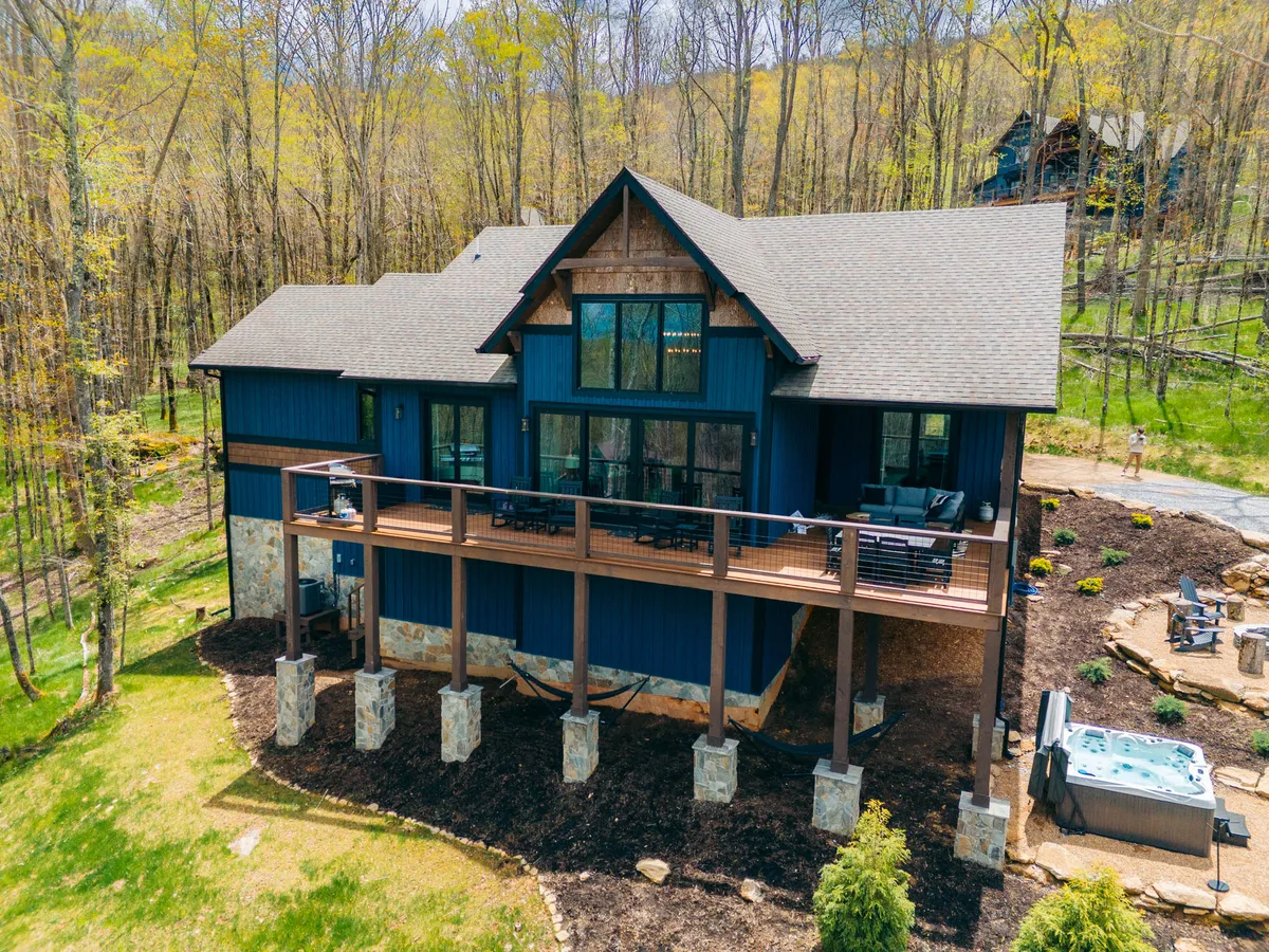 A wide shot of the main deck, showcasing rocking chairs and a dining table set against the backdrop of the home's blue and wood exterior and surrounded by lush trees. The image captures both the hot tub and seating areas, suggesting opportunities for relaxation and outdoor dining while immersed in nature.