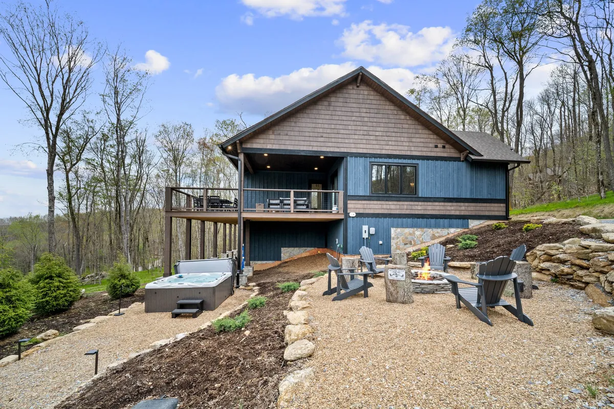 The image showcases the modern cabin architecture of the home with a focus on the outdoor hot tub, fire pit, and a comfortable seating area. The natural surroundings and evening lighting evoke a sense of tranquility and togetherness.