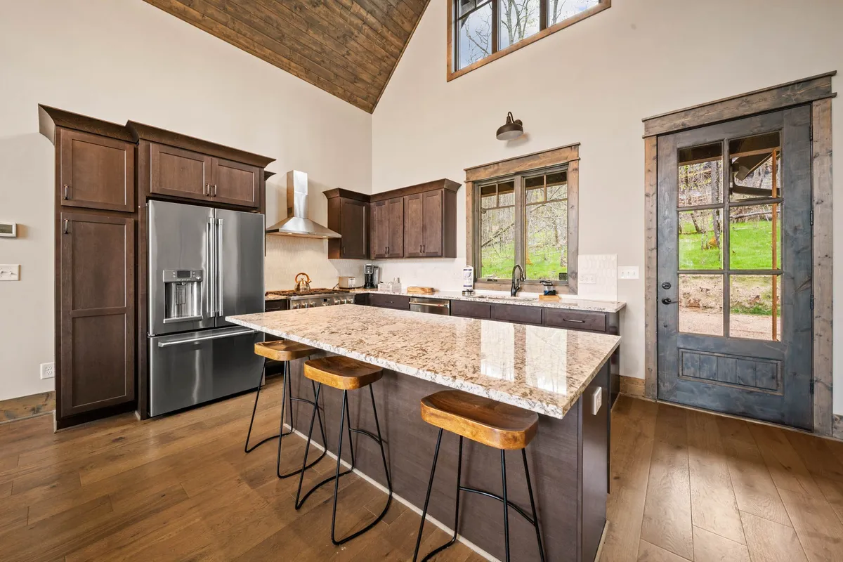 A bright, open-concept kitchen with granite countertops, a spacious island with barstool seating, rustic cabinetry, and large windows filling the space with natural light. The layout encourages family cooking and gathering, while the island offers a welcoming spot for casual meals or conversations.