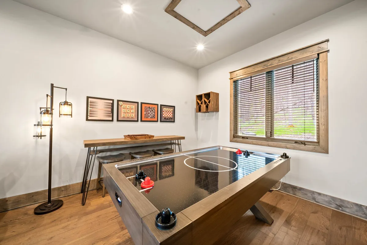 A bright, inviting game room setting with an air hockey table, decorative wall art, a window with blinds letting in natural light, and wooden furniture. The arrangement is conducive to social gatherings and fun activities, emphasizing the playful aspect of the space.
