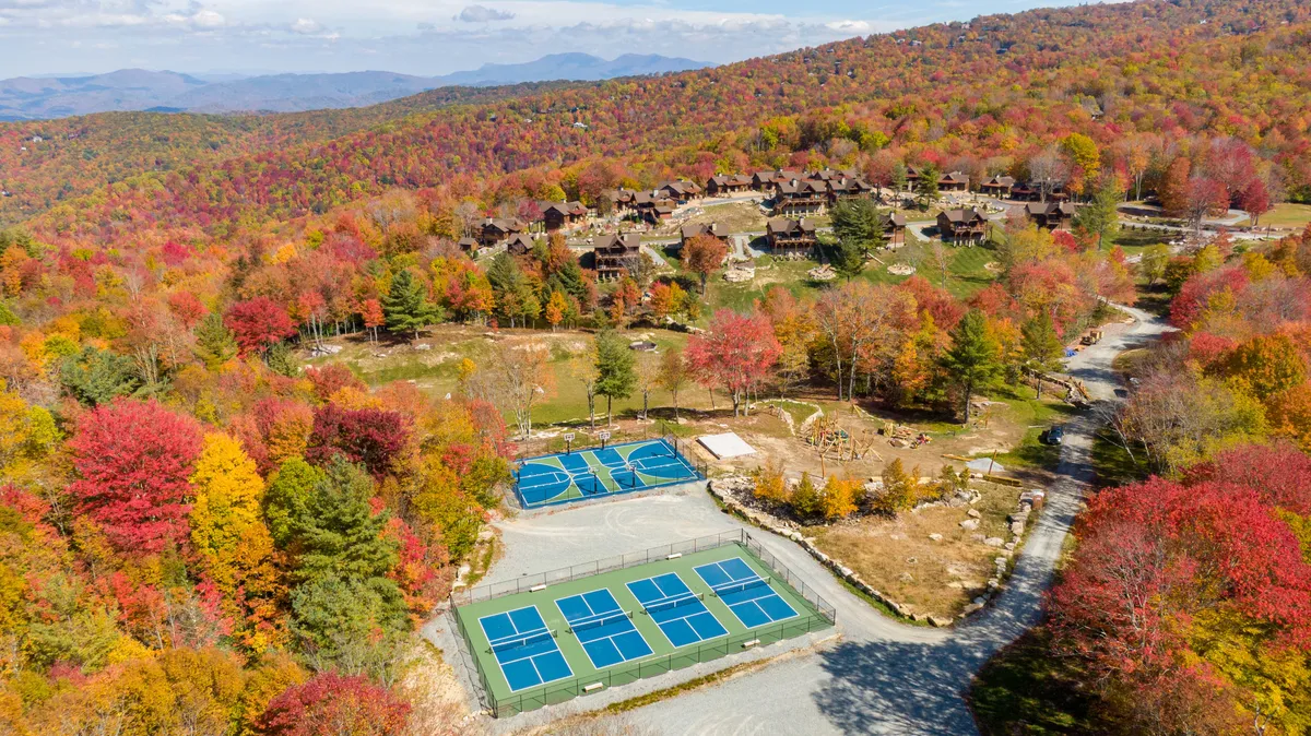 An exterior shot featuring tennis courts surrounded by colorful autumn foliage, wooden cabins, and a striking mountain backdrop. Families can be seen playing tennis and enjoying the beautiful natural setting, emphasizing opportunities for outdoor recreation and bonding.
