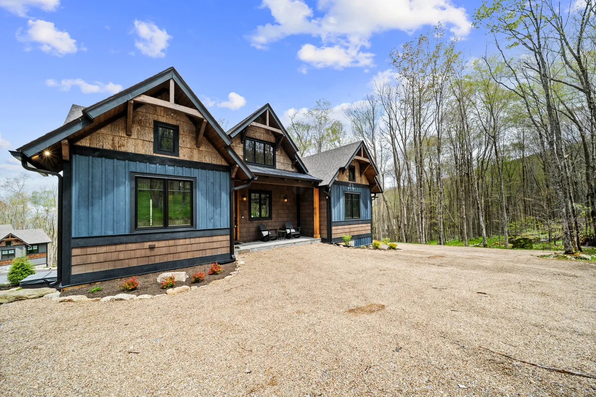 This image presents the unique architectural design of the house set amidst a lush, wooded environment. The gravel driveway leads to a welcoming porch, while the natural setting with dense trees highlights the home's connection to the outdoors and the changing seasons.